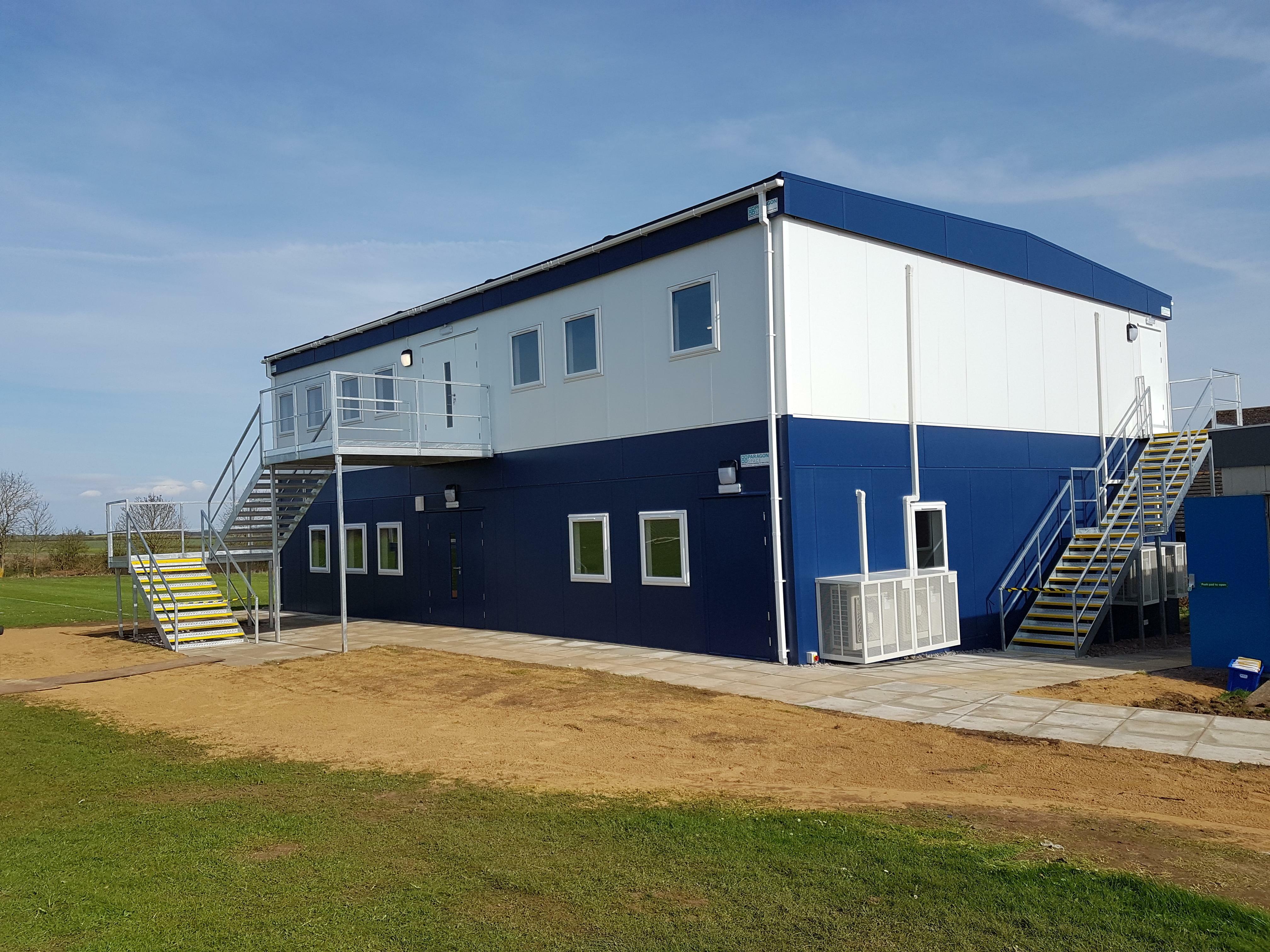 A two-story modular building features exterior metal staircases on both sides. It is situated on a grassy field with a newly laid sidewalk. The sky is clear.