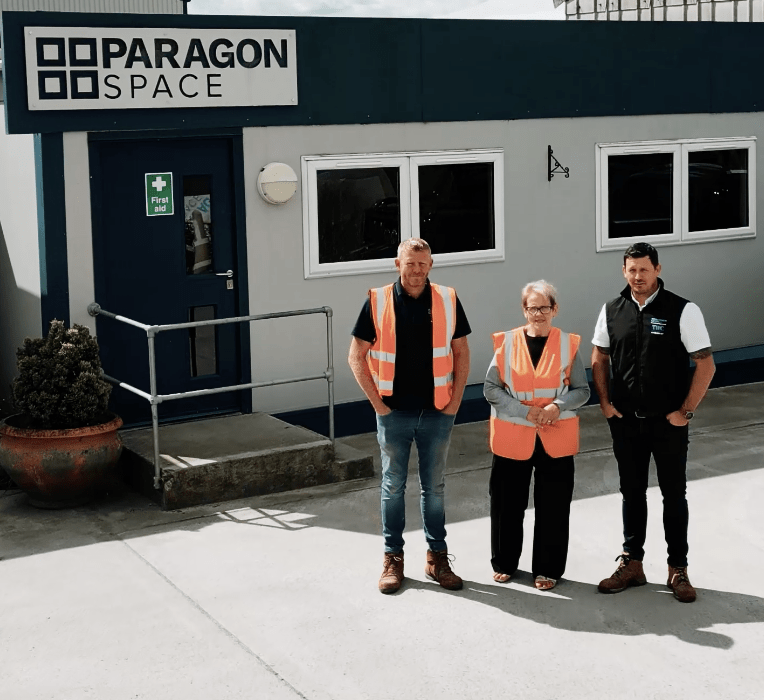 Three people stand in front of a grey portable cabin labeled 
