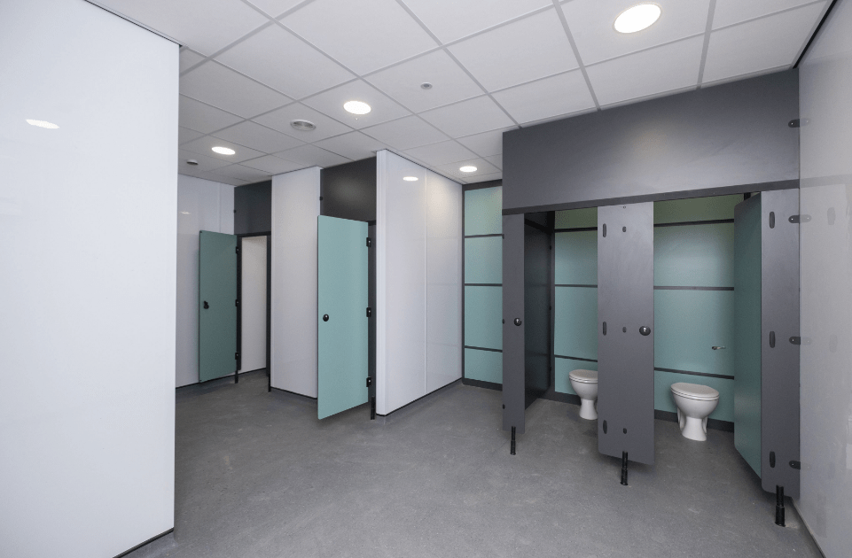 Toilet stalls with open doors in a public restroom, revealing white porcelain toilets, under fluorescent ceiling lights in a clean, modern space with teal-colored partitions and gray flooring.