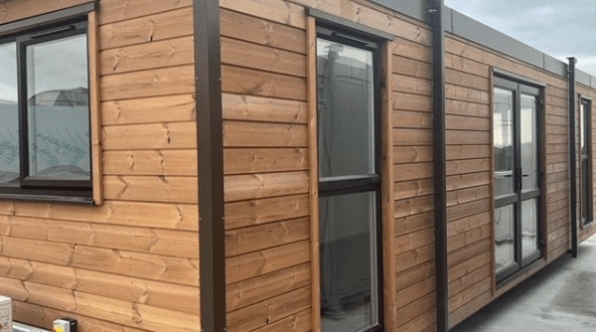 Wooden modular cabin featuring several windows along its outer walls, placed on a concrete surface under an overcast sky.