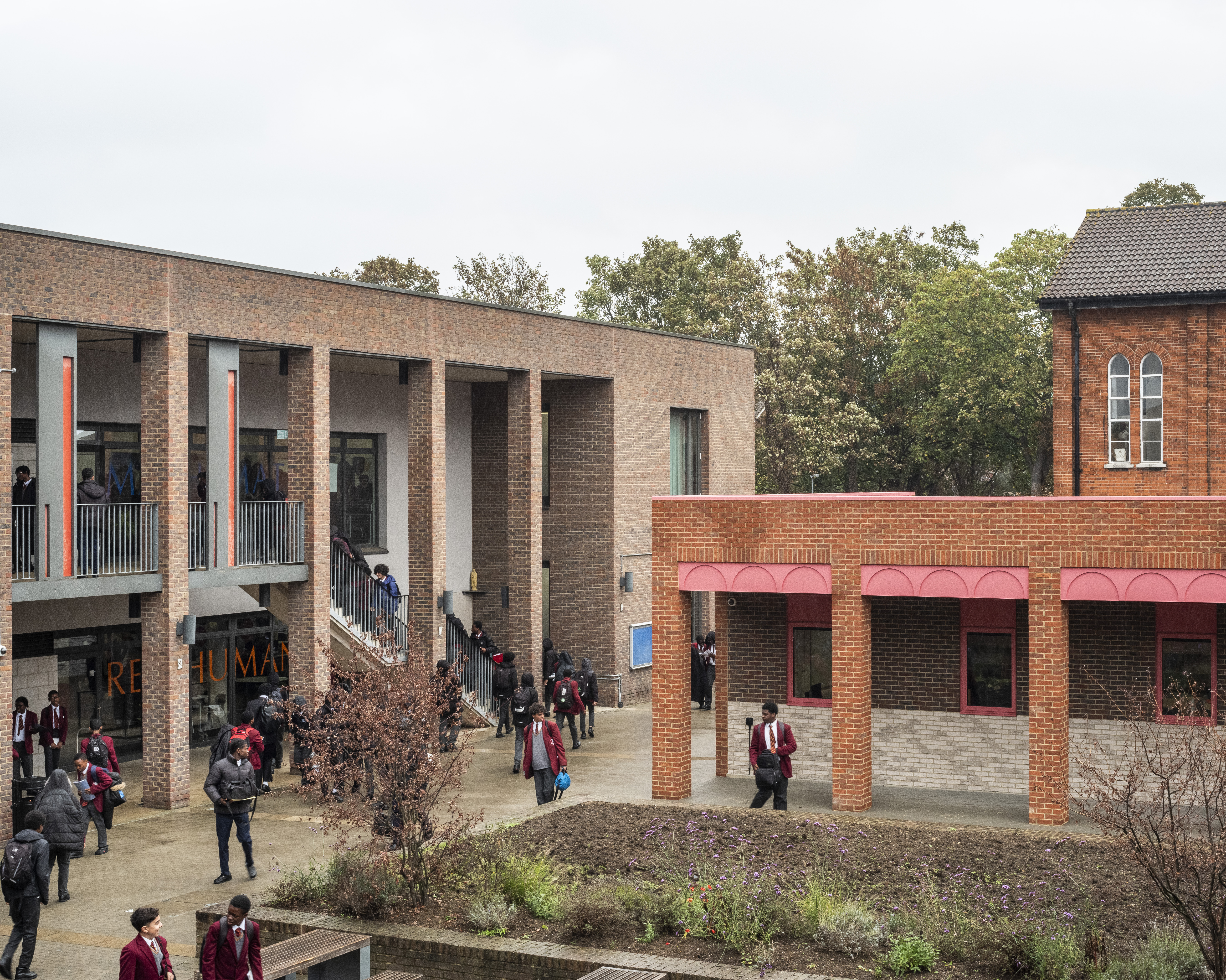 Students, many in red uniforms, are walking and gathering around a multi-story brick school building with red accents, featuring exterior staircases and corridors, surrounded by trees and a garden bed.
