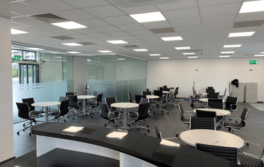 Several round tables with black office chairs arranged in a brightly lit, modern office space featuring floor-to-ceiling glass walls and a curved reception counter. An exit sign is visible.