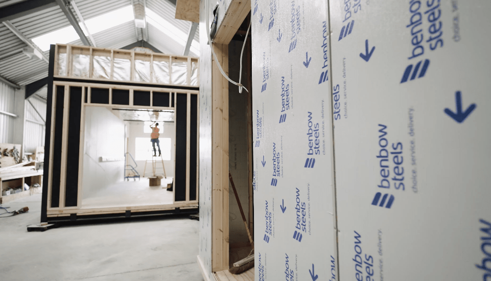 A partially constructed room with wall insulation takes center stage. A worker stands on a ladder inside, installing electrical wiring. Surrounding context includes construction materials and tools in a metal-framed warehouse. Text on wall insulation reads, 