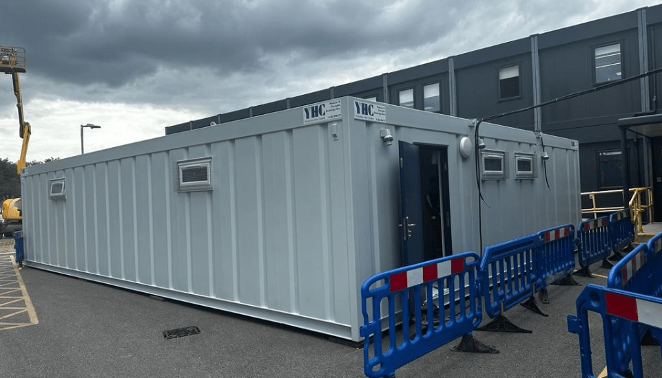 A large white temporary modular building with a partially open door, positioned in an industrial parking lot, surrounded by blue safety barriers. The text on the building reads 