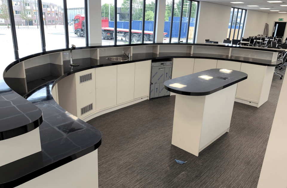 A curved countertop with sinks and storage cabinets in a bright room with large windows. In the background, a red truck and blue containers are visible outside.