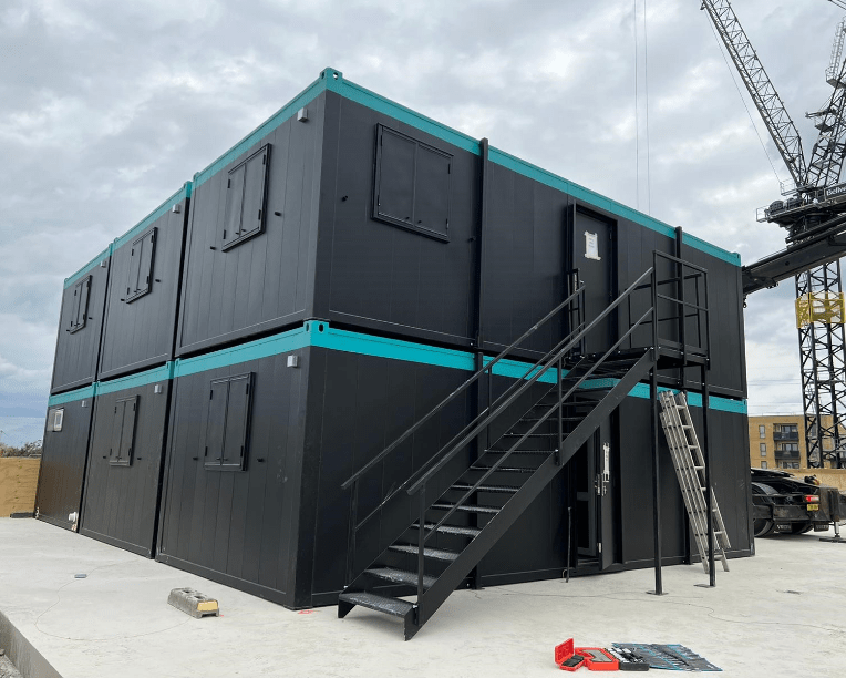 A two-story black modular building with blue trim has an external staircase and ladder. It is situated on a concrete platform, and a crane is visible in the background.