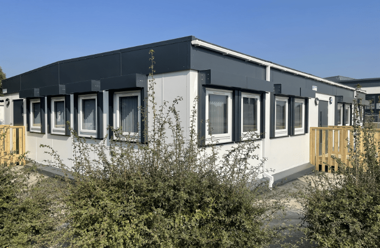 Rectangular modular building with several windows, surrounded by shrubs and bushes, under a clear blue sky. Wooden railings are by the entrance. No text is visible.