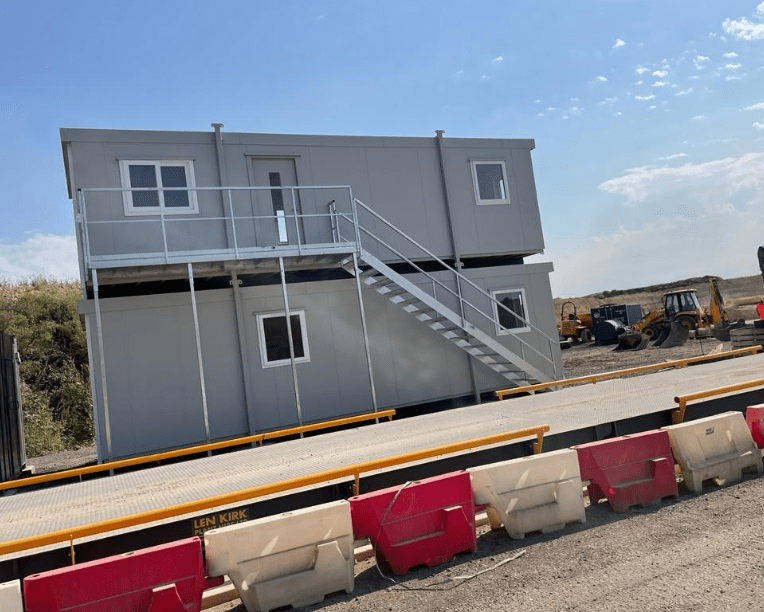 A two-story portable building with exterior stairs stands on a construction site, surrounded by equipment and safety barriers labeled 